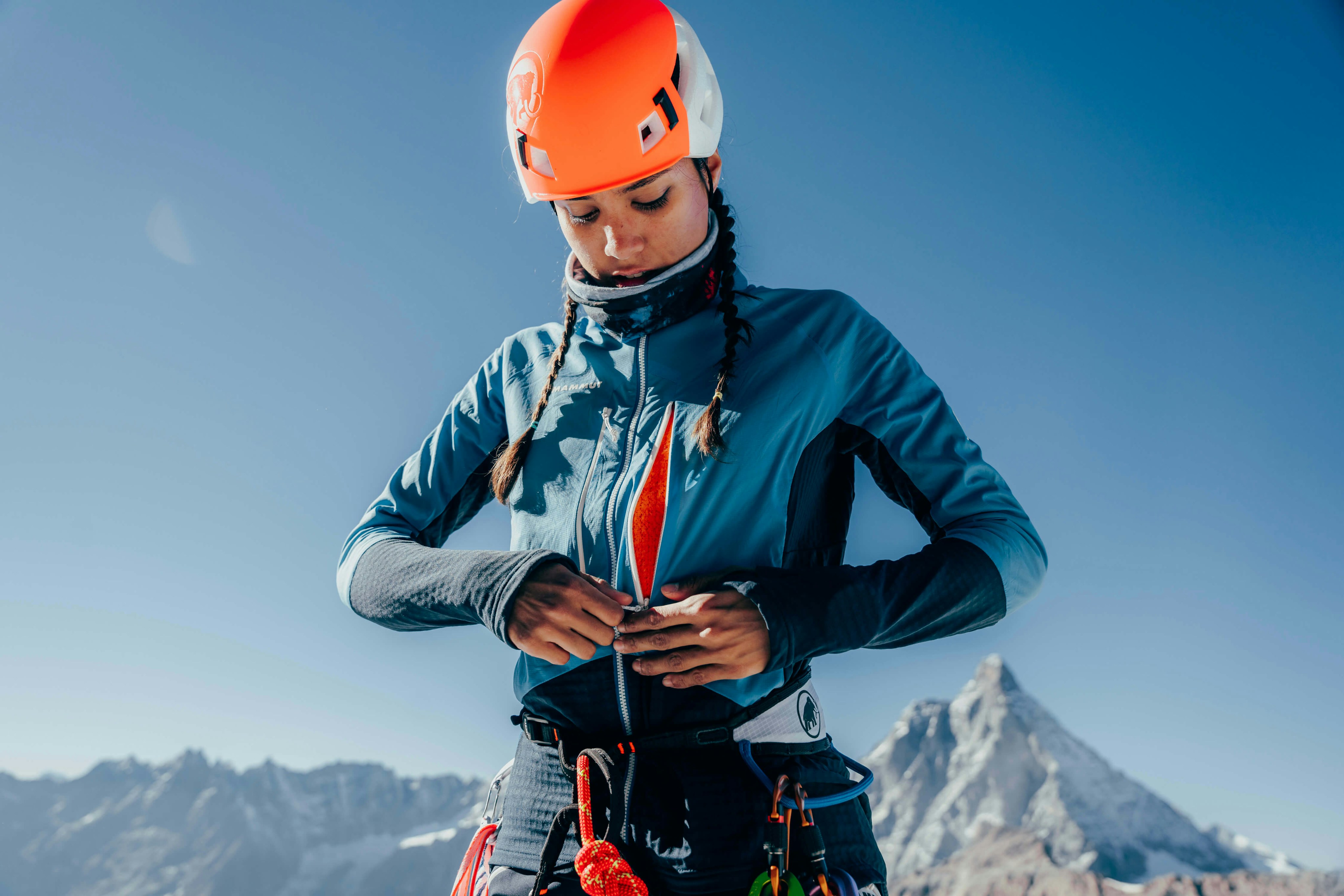 A mountain climber wearing a blue Mammut jacket and an orange helmet is adjusting their climbing gear. A snowy peak rises dramatically in the background.