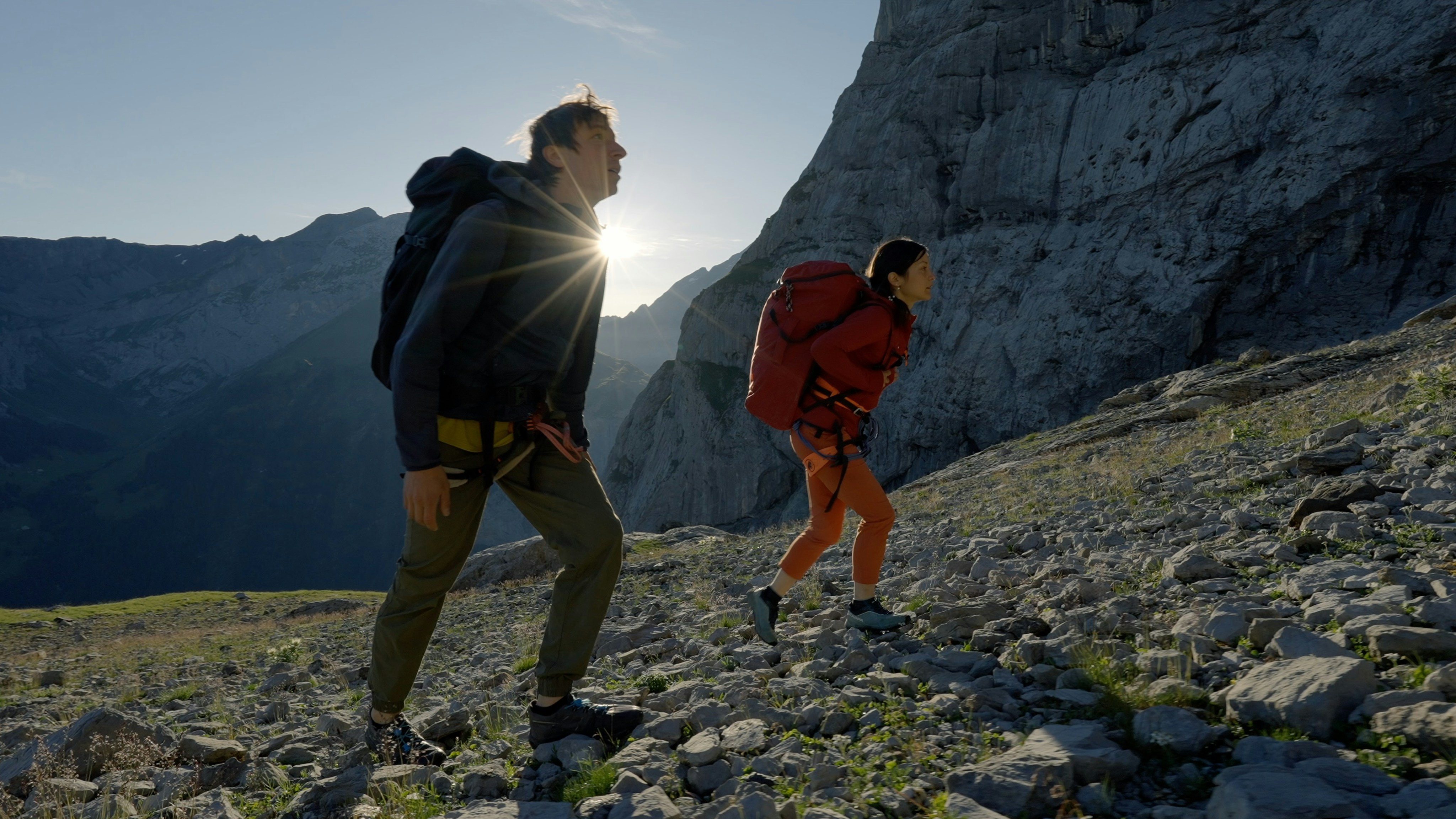 Katherine Choong & Jim Zimmermann, two Mammut-clad hikers with large backpacks ascending a rugged mountain slope at sunrise.