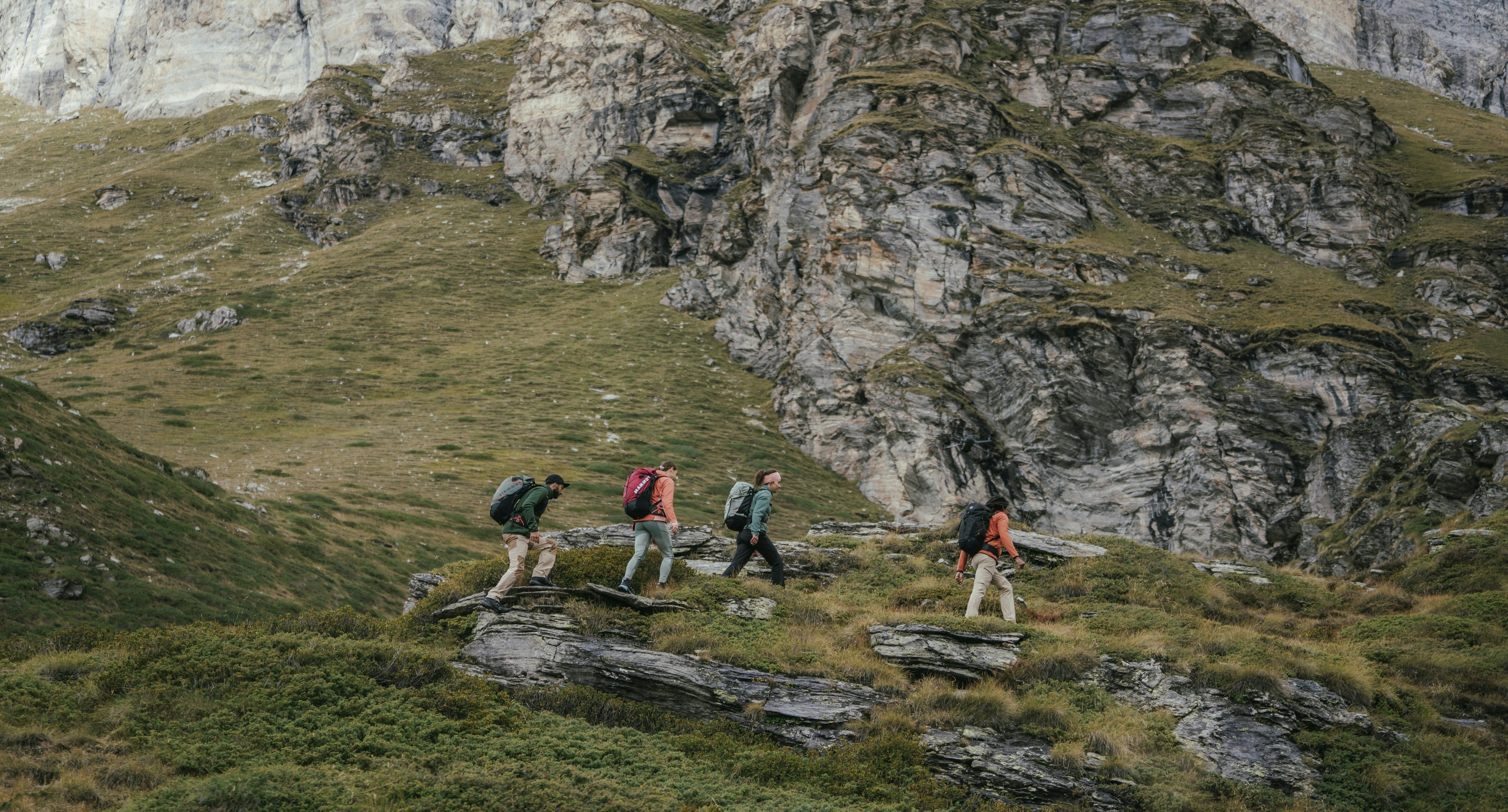 Four Mammut-clad hikers with backpacks trek in unison along a rocky, grassy trail set against the majestic backdrop of steep, rugged mountains. The vibrant landscape features lush green patches of shrubs and striking stratified cliff faces.