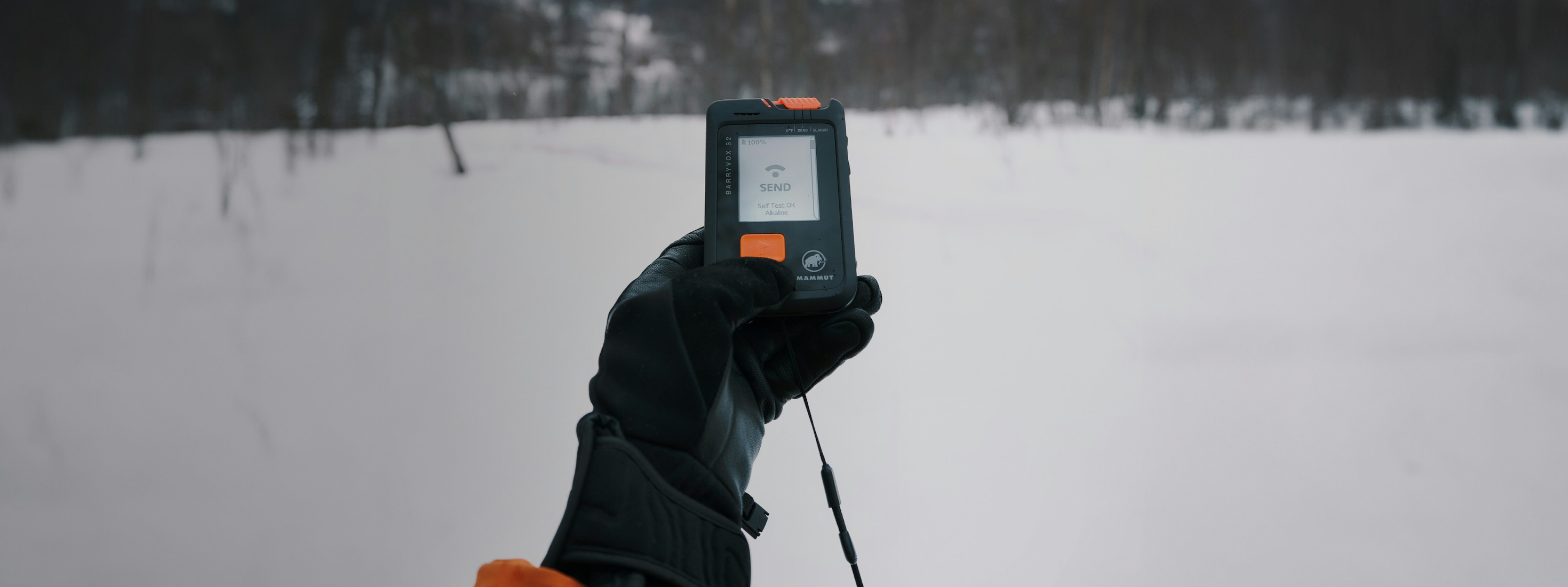 Gloved hand holding a Mammut avalanche transceiver displaying "SEND" on the screen, with a snowy mountain landscape in the background.