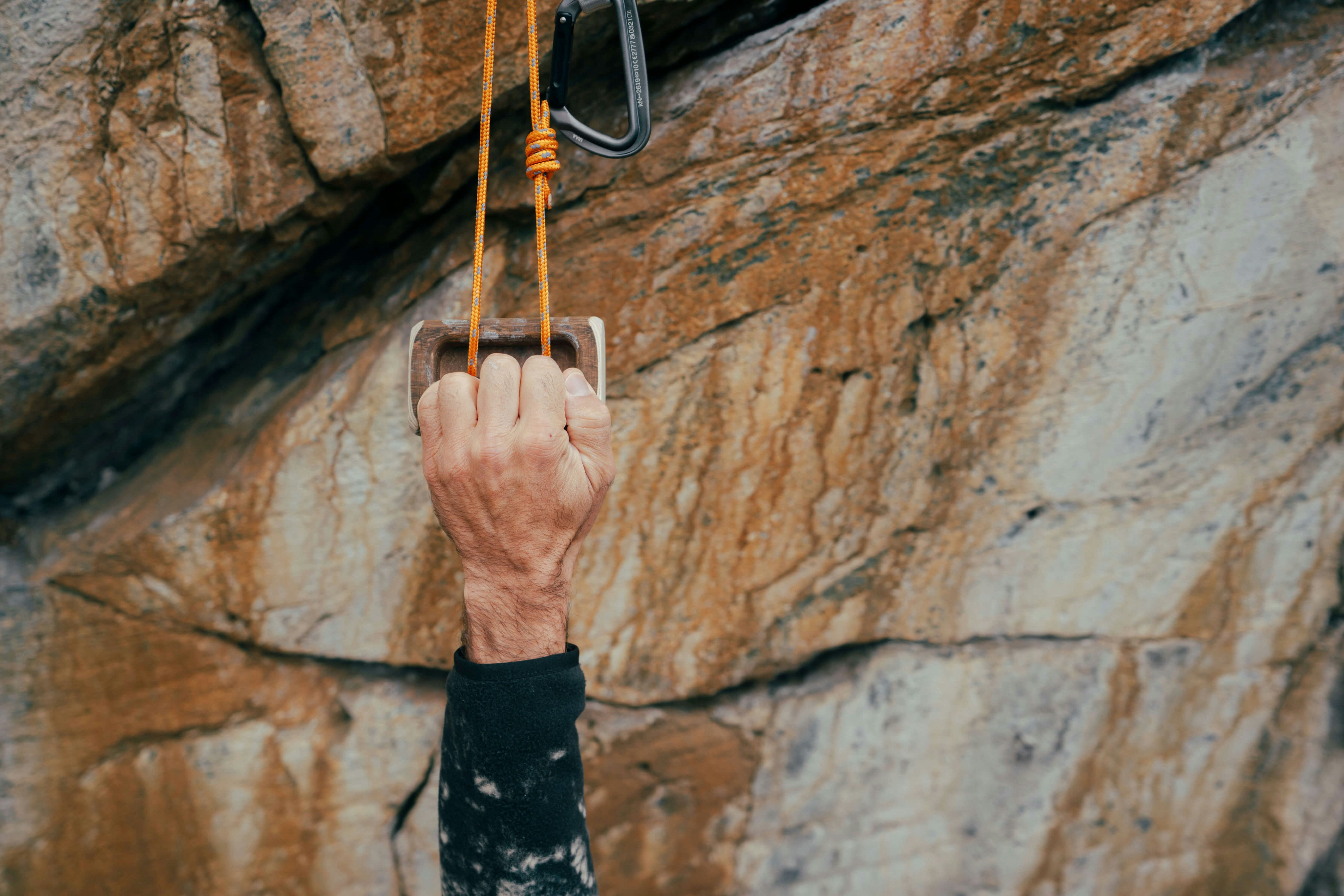 A hand gripping a climbing hold on a rocky surface, with a Mammut rope securely clipped to a carabiner above.