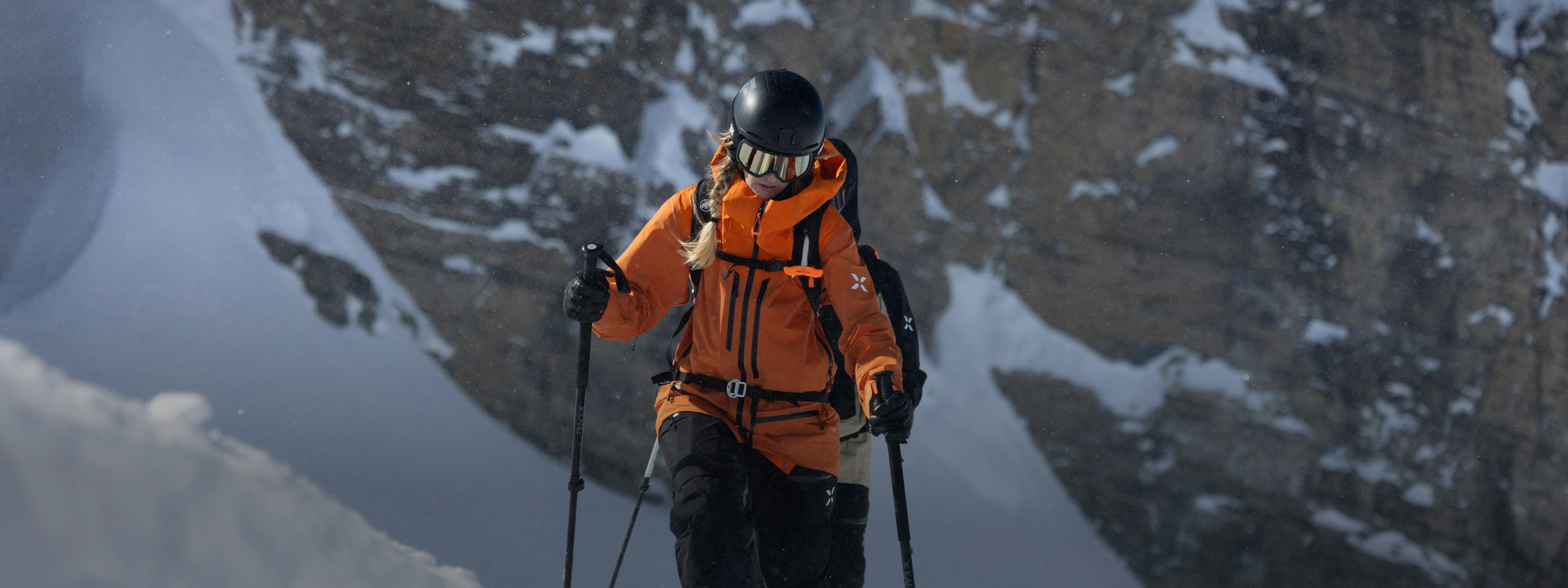 Sanne. a skier wearing a Mammut orange jacket and helmet navigates down a snowy mountain with rugged rocky cliffs in the background.