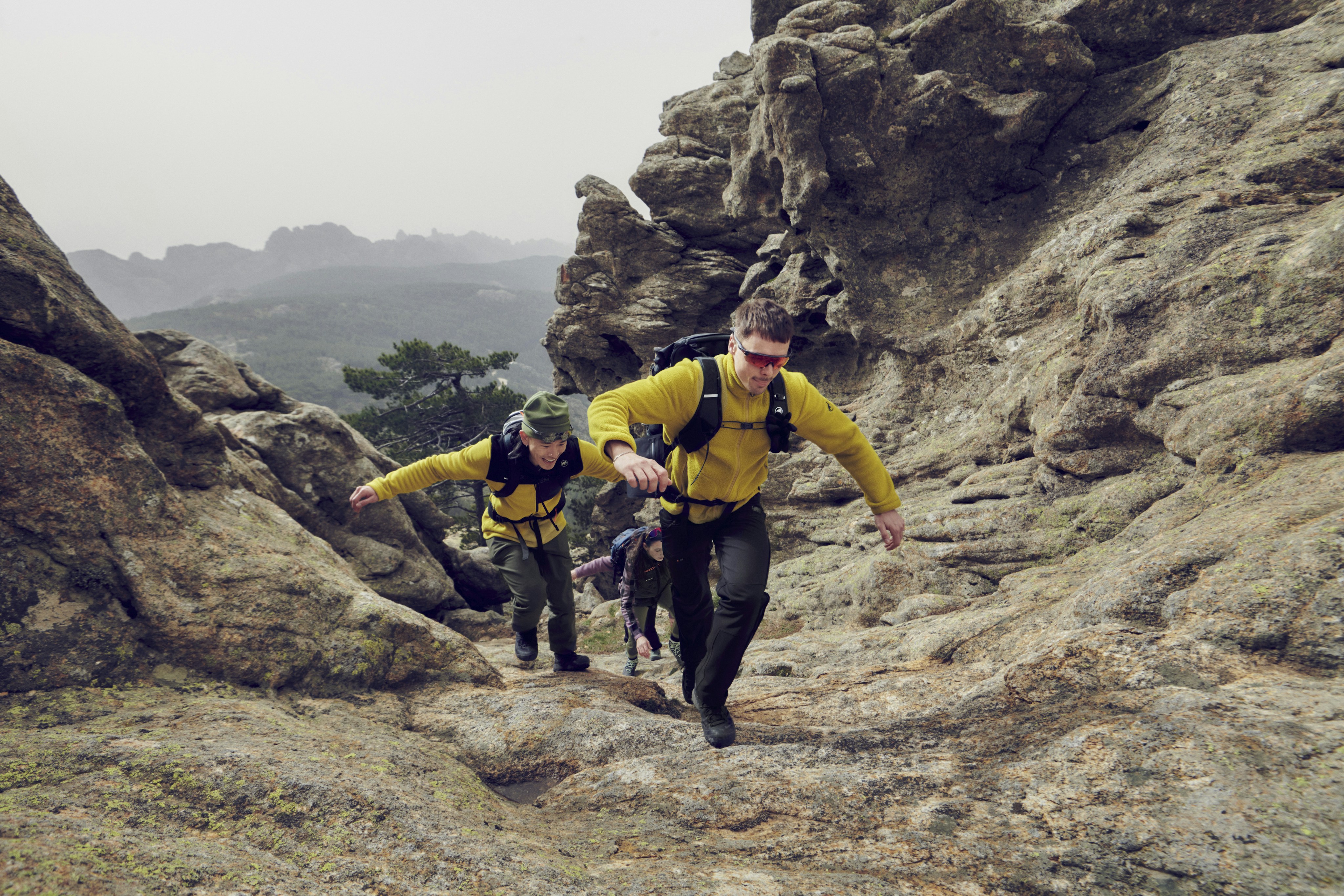 Two hikers in Mammut yellow jackets climb a rocky trail in a rugged mountain landscape.