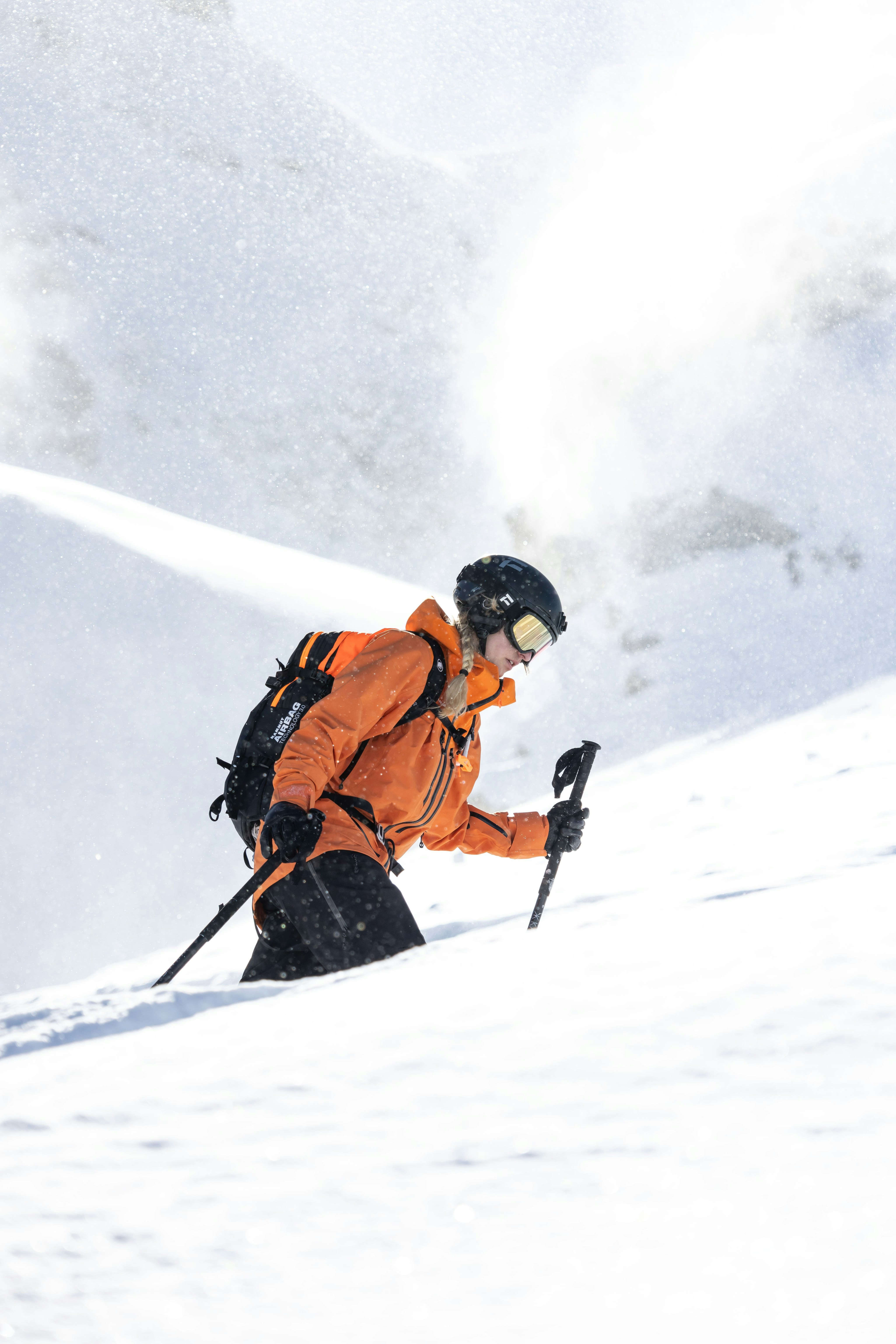 Person wearing a Mammut orange jacket skiing expertly through deep powder snow on a bright, sunny day.