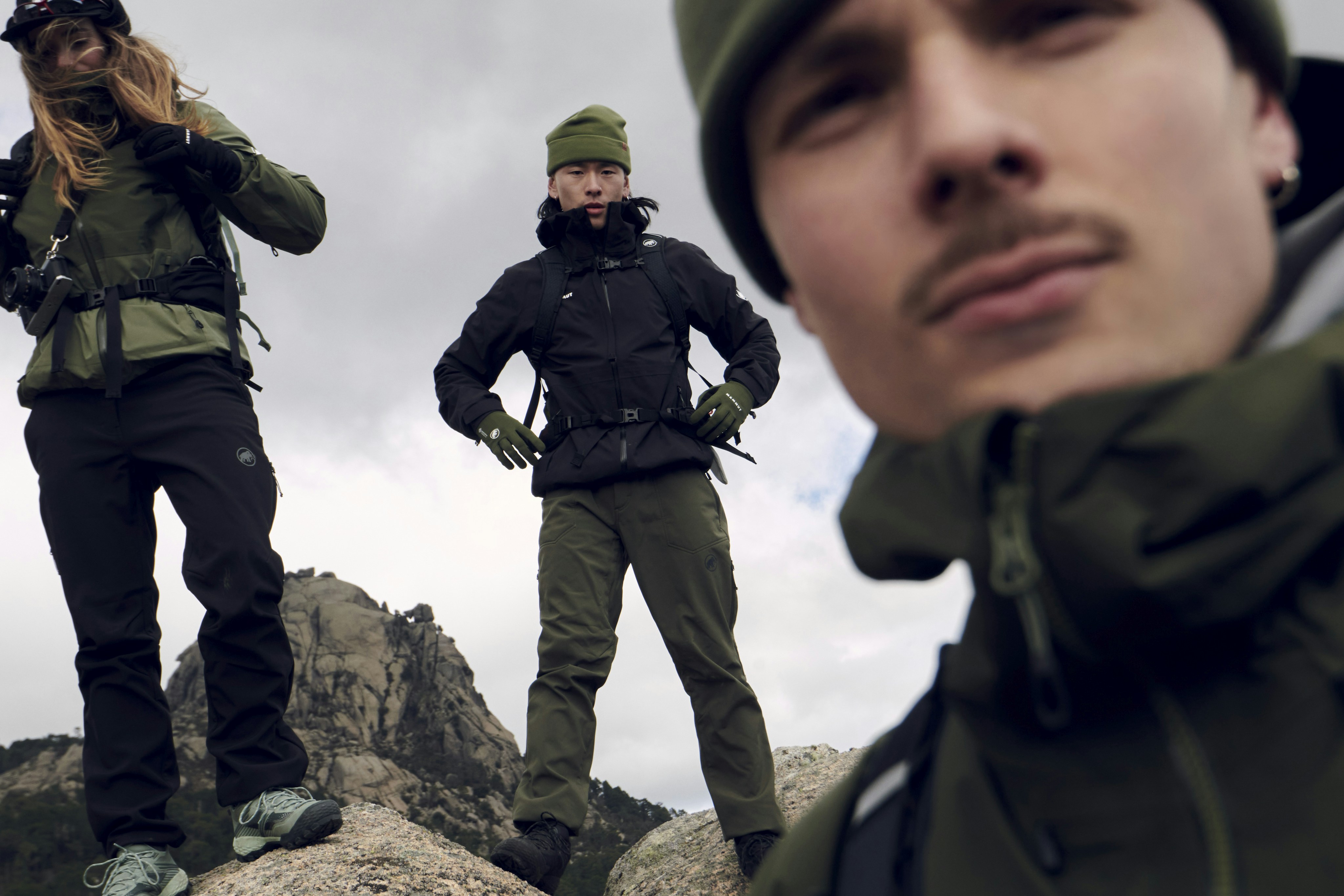 Three adventurers in Mammut outdoor gear stand on a rocky mountain, with one person close to the camera and two others positioned in the background.