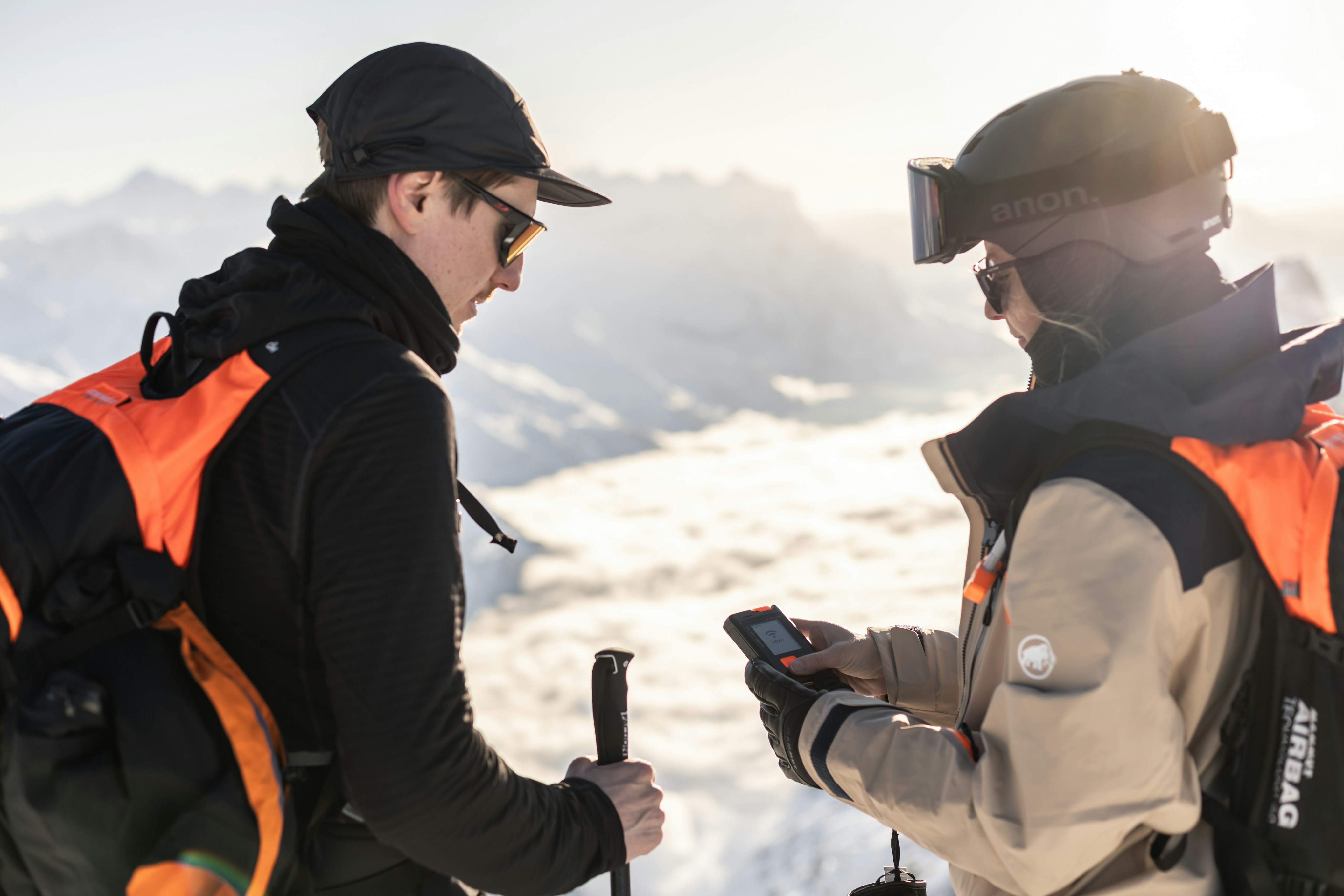 Two hikers in Mammut winter gear and sunglasses stand on a snowy mountain; one is holding Mammut trekking poles, while the other checks a navigation device.