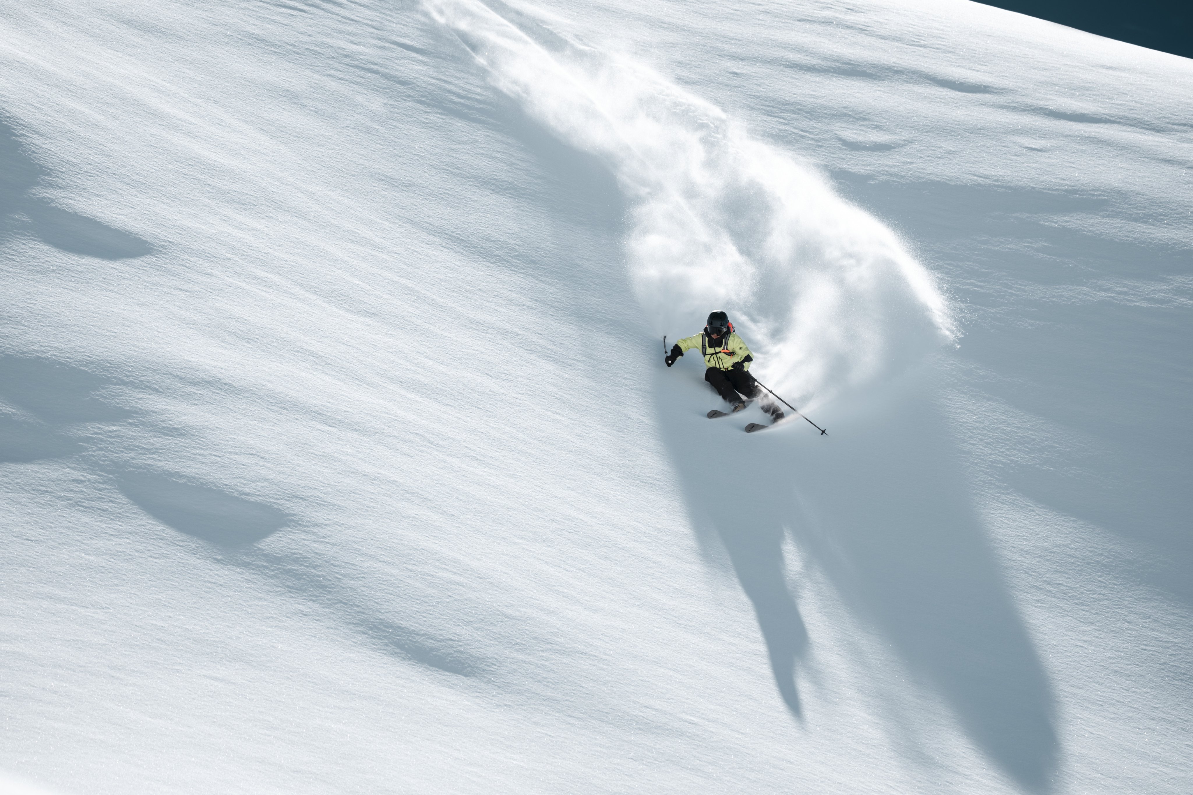 Sanne Mona wearing a Mammut orange jacket and black helmet effortlessly glides through pristine snowy terrain, leaving a trail of sparkling powder behind.