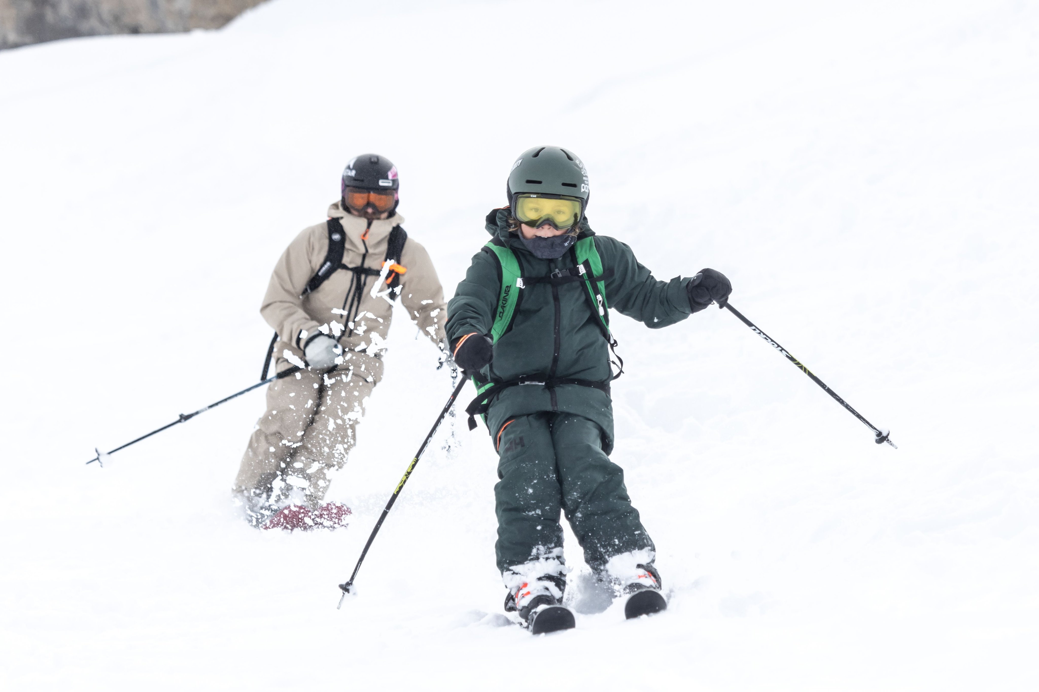 Whisky and his son in Mammut winter gear expertly carving down a snow-covered slope, with powdery snow dramatically flying up around them.