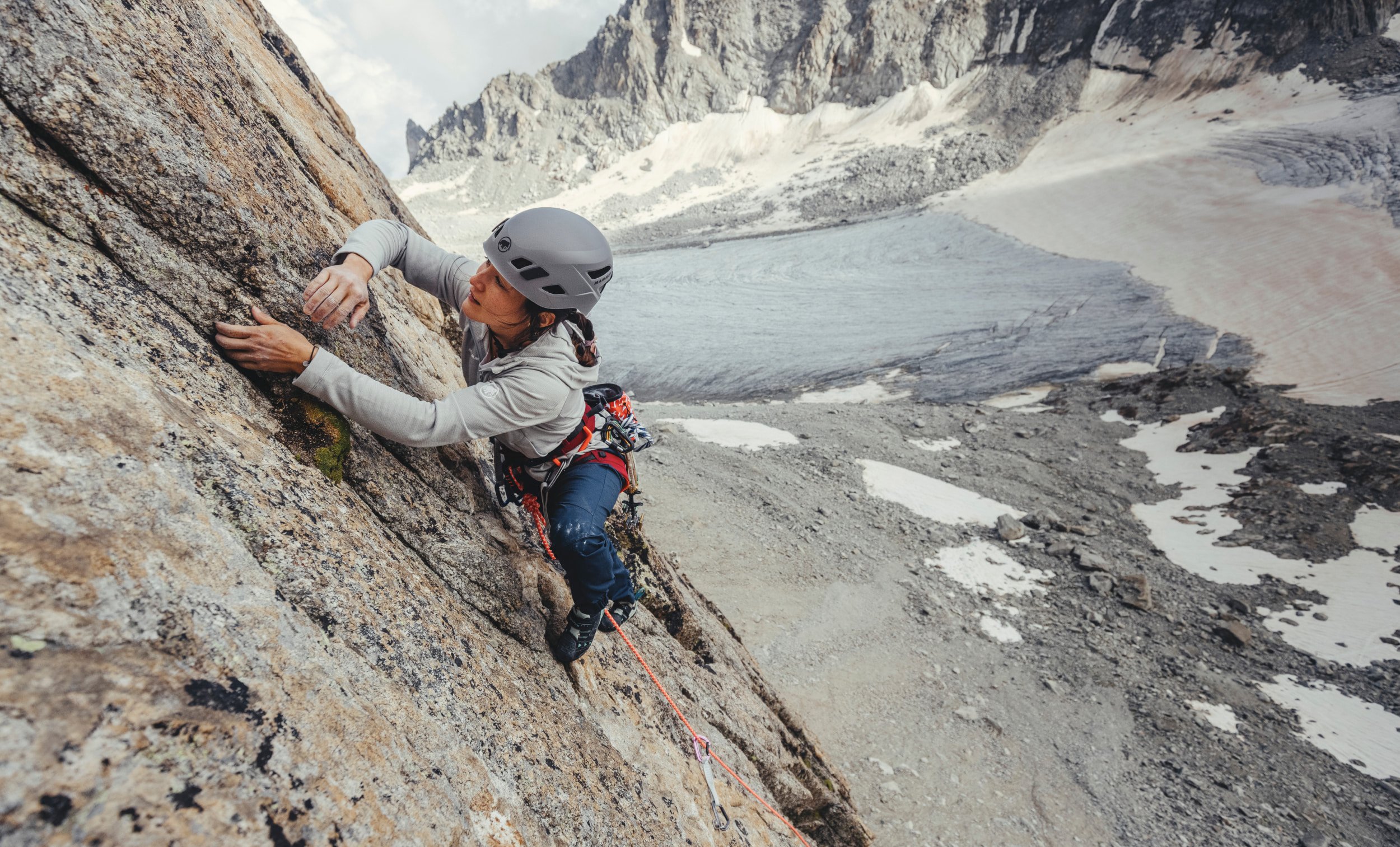 Person climbing a steep rock face with snowy mountain peaks in the background, wearing Mammut helmet and climbing gear.
