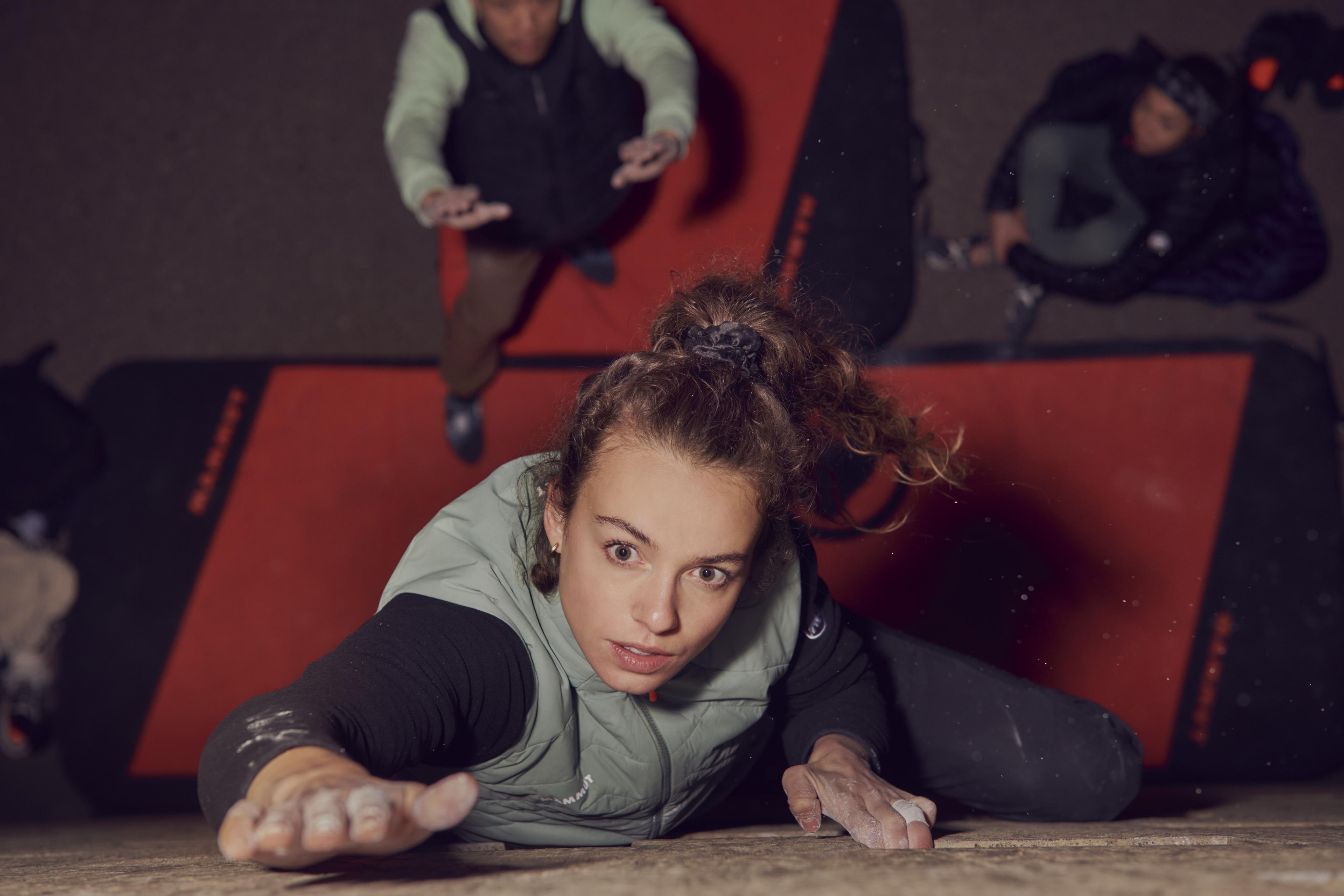 A woman climbs a rock wall wearing Mammut climbing gear, with two people below on crash mats prepared to catch her if she falls.