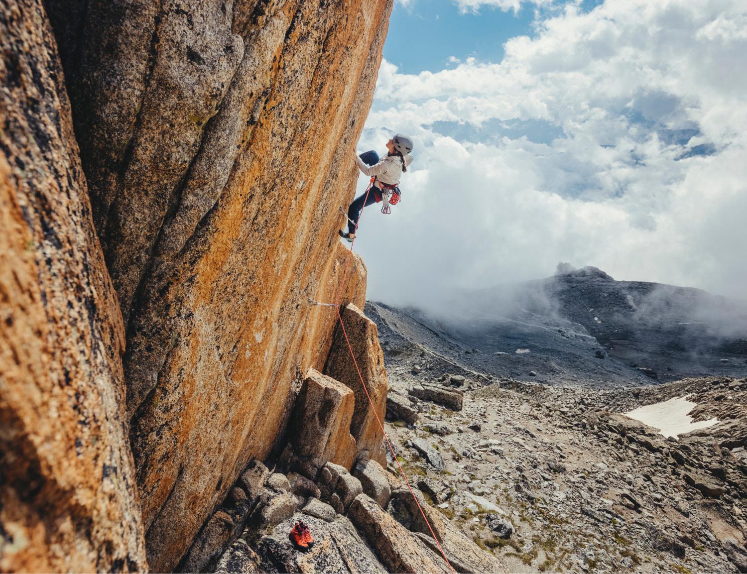 Person rock climbing a steep mountain wearing Mammut gear with clouds gathering in the background on a rugged rocky landscape.