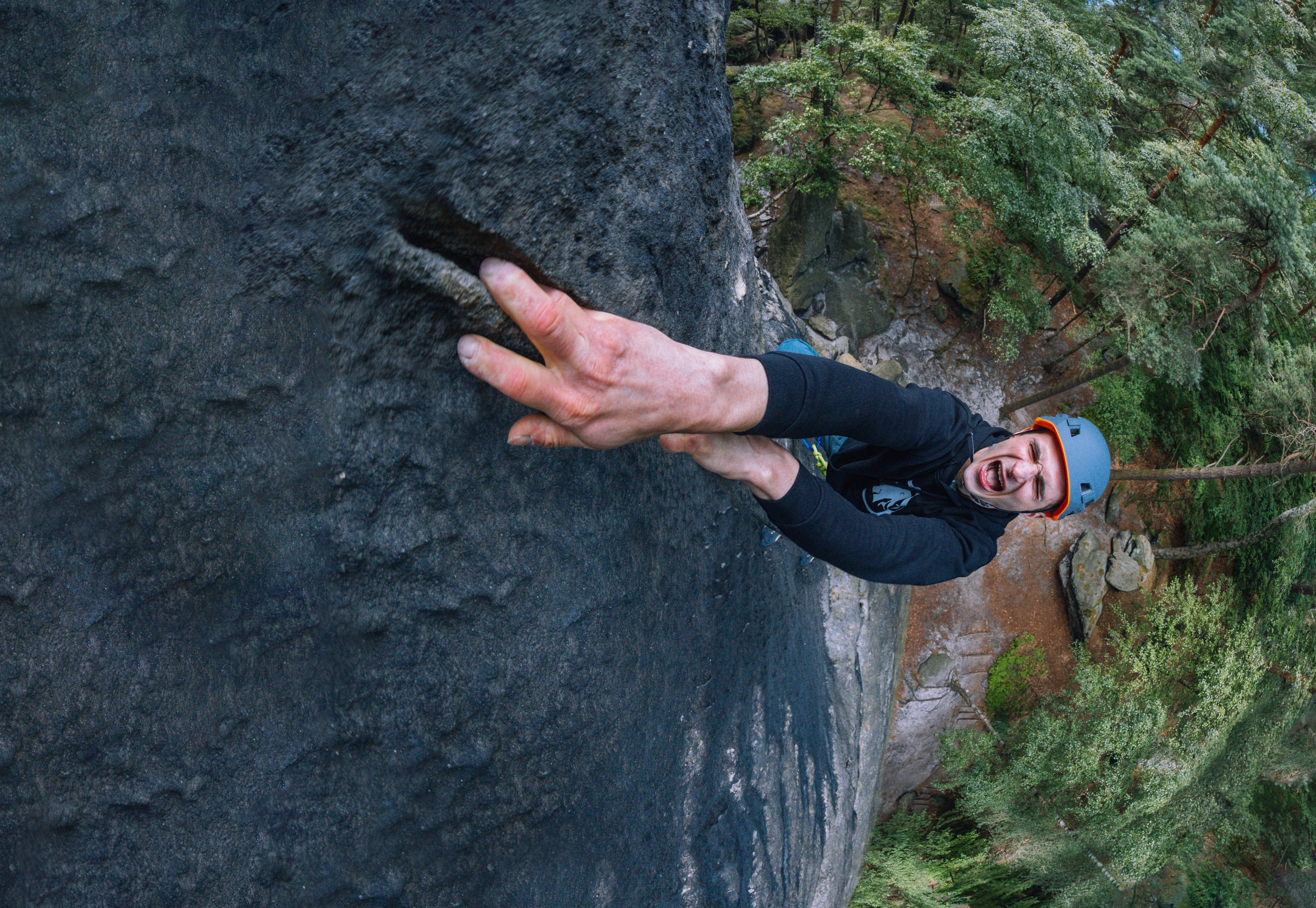 Man wearing a Mammut helmet climbs a steep rock face, gripping a crevice with his fingers; dense forest stretches below.