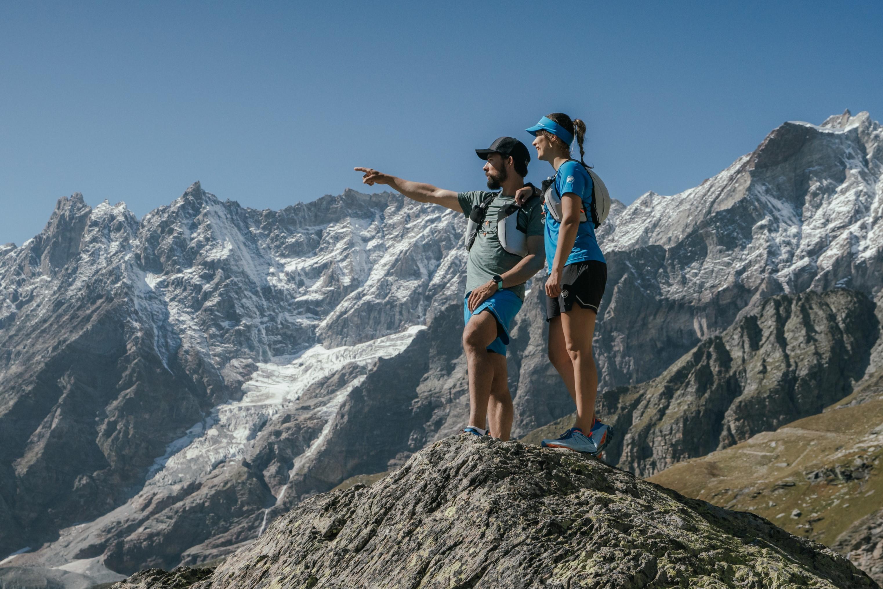 Two hikers outfitted in blue and grey Mammut gear standing on a rocky peak, with stunning snowy mountains in the background under a clear sky.