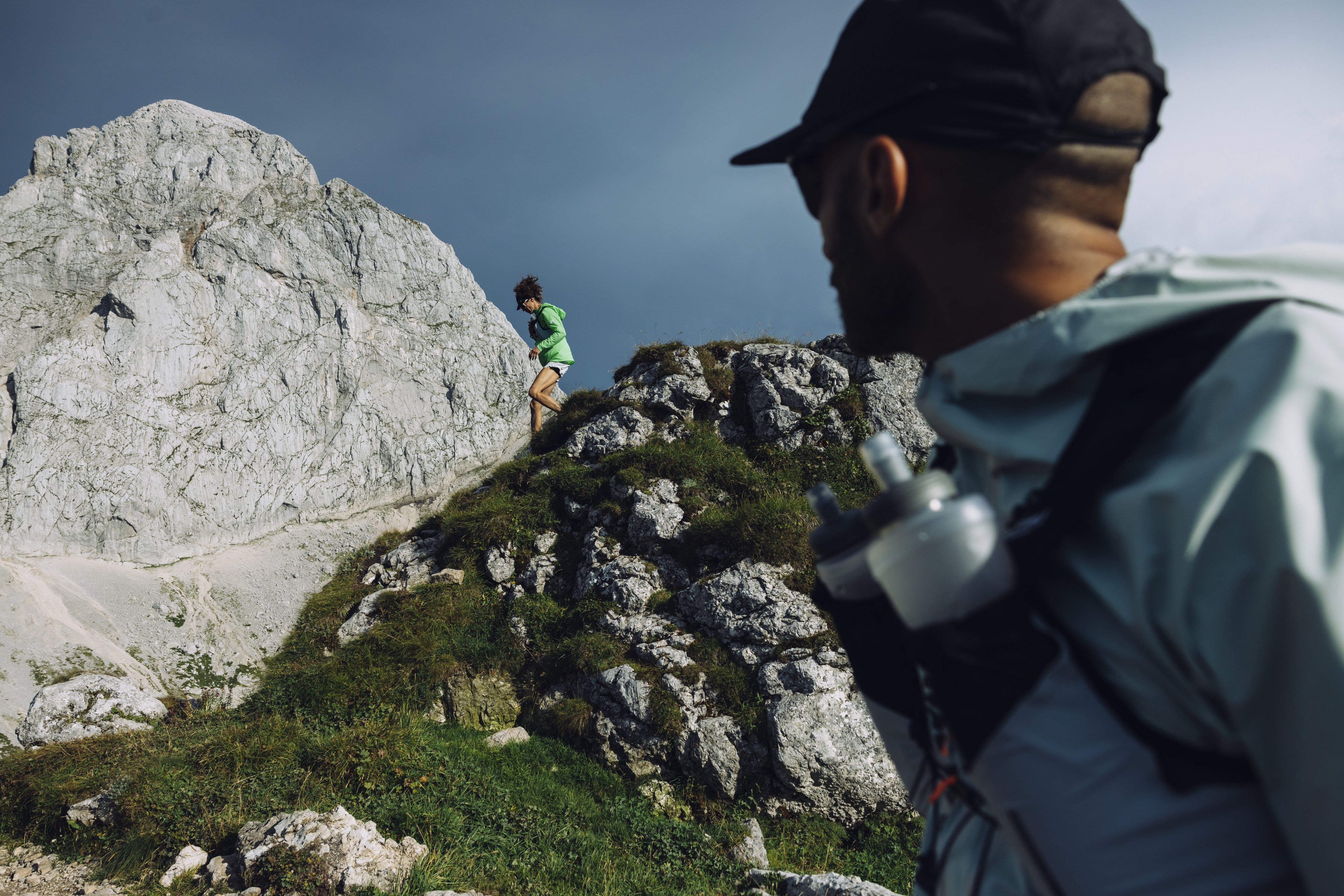 Mammut-clad hiker ascends rocky hill in the distance, while another person in Mammut outdoor gear watches from the foreground.