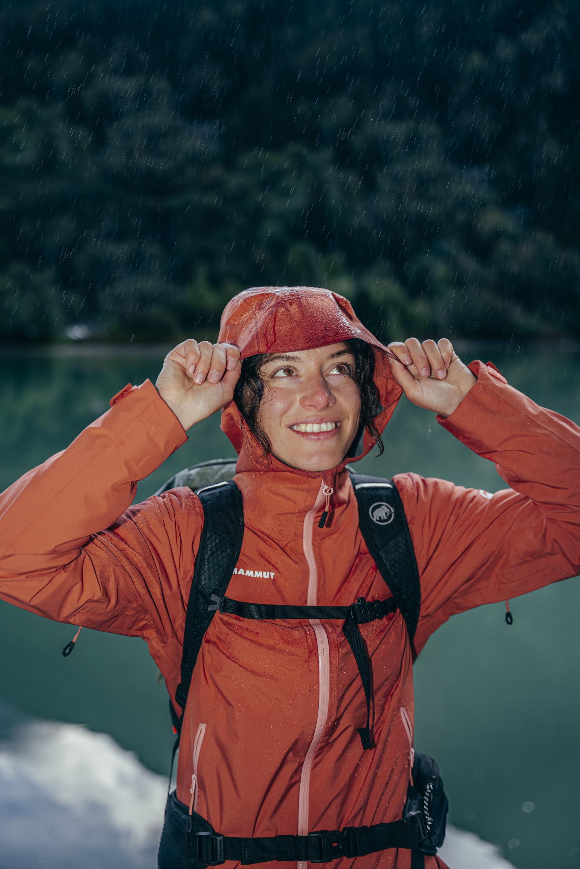 Person smiling in a Mammut orange rain jacket, adjusting the hood outdoors near a scenic lake and lush greenery in the background.