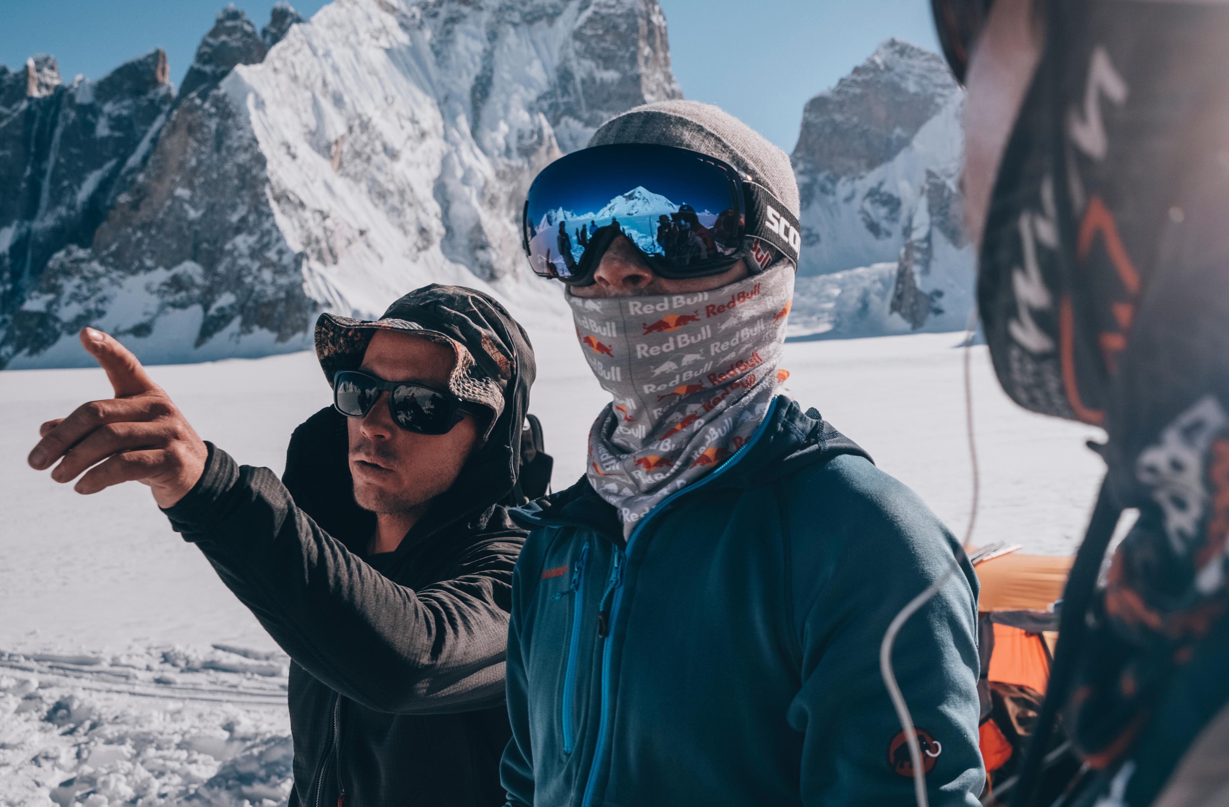Two climbers in Mammut cold-weather gear on a snowy mountain, one pointing ahead, both protected with sunglasses and face coverings.
