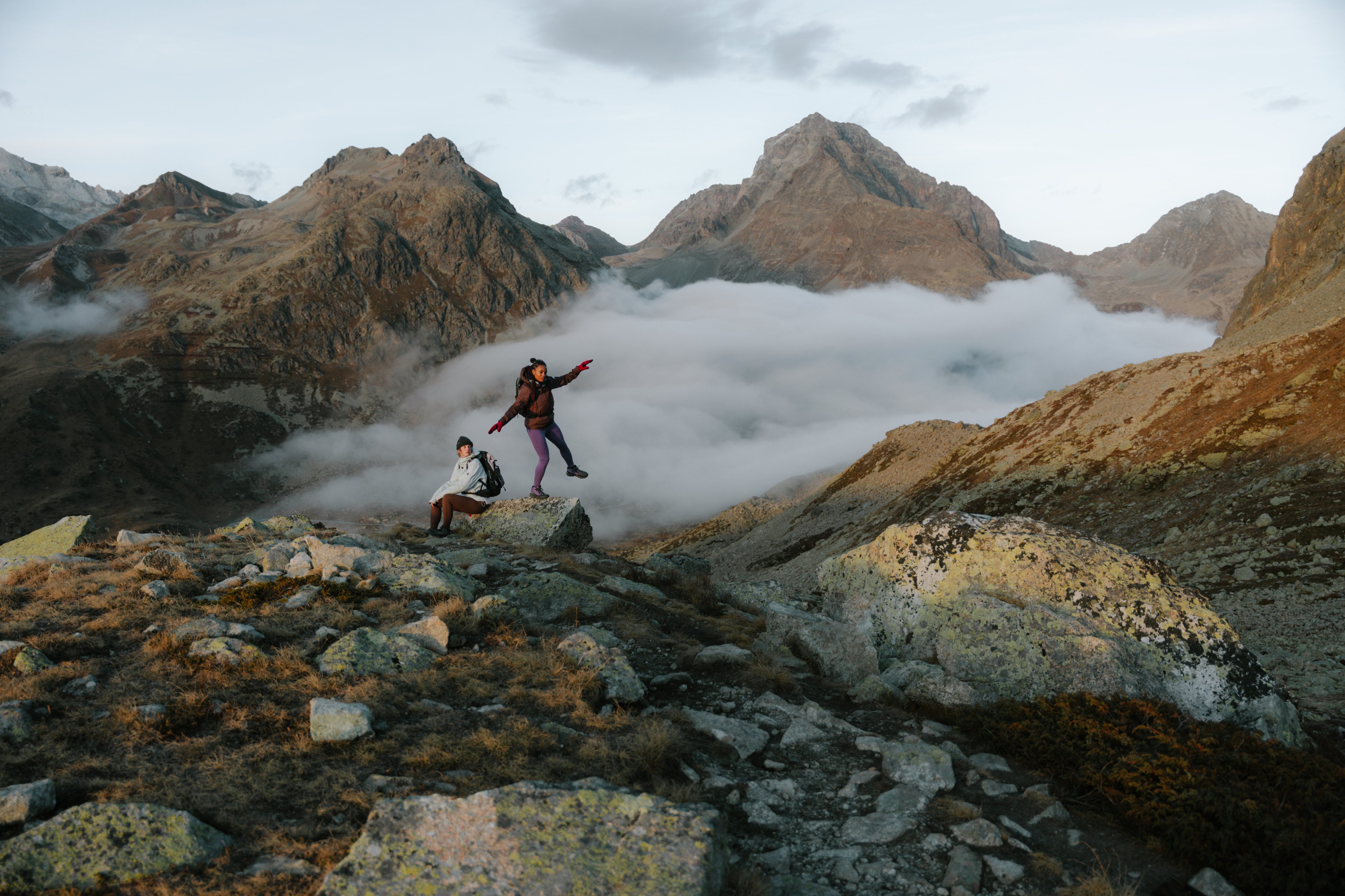 Two hikers in Mammut gear on rocky mountain terrain, one mid-jump, with clouds and alpine peaks in the background.