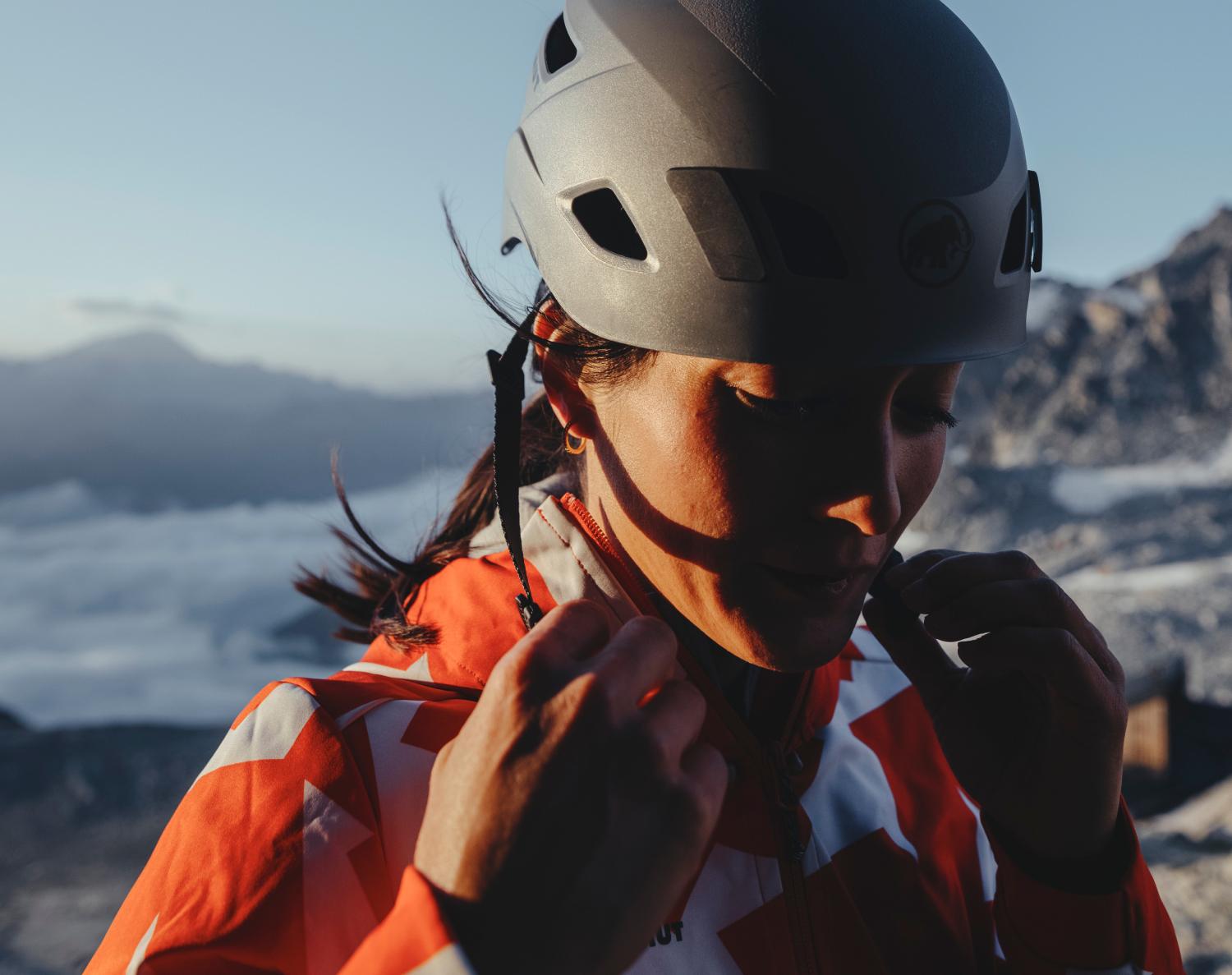 Person wearing a Mammut helmet and adjusting their Mammut jacket, with a breathtaking mountainous landscape in the background.