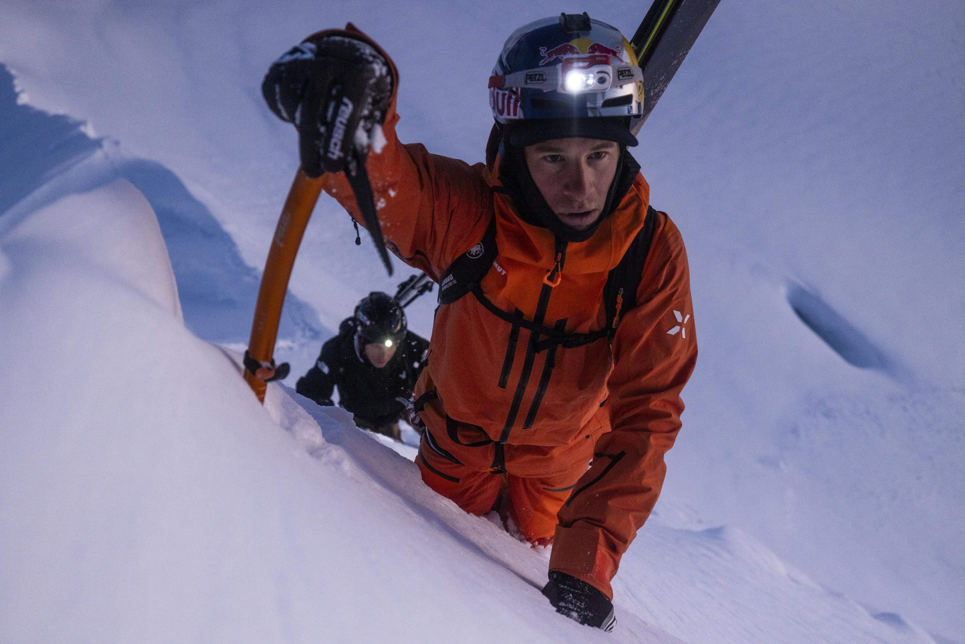 Two climbers in bright orange Mammut gear ascend a challenging snowy slope, one wielding an ice axe and both equipped with headlamps for visibility.