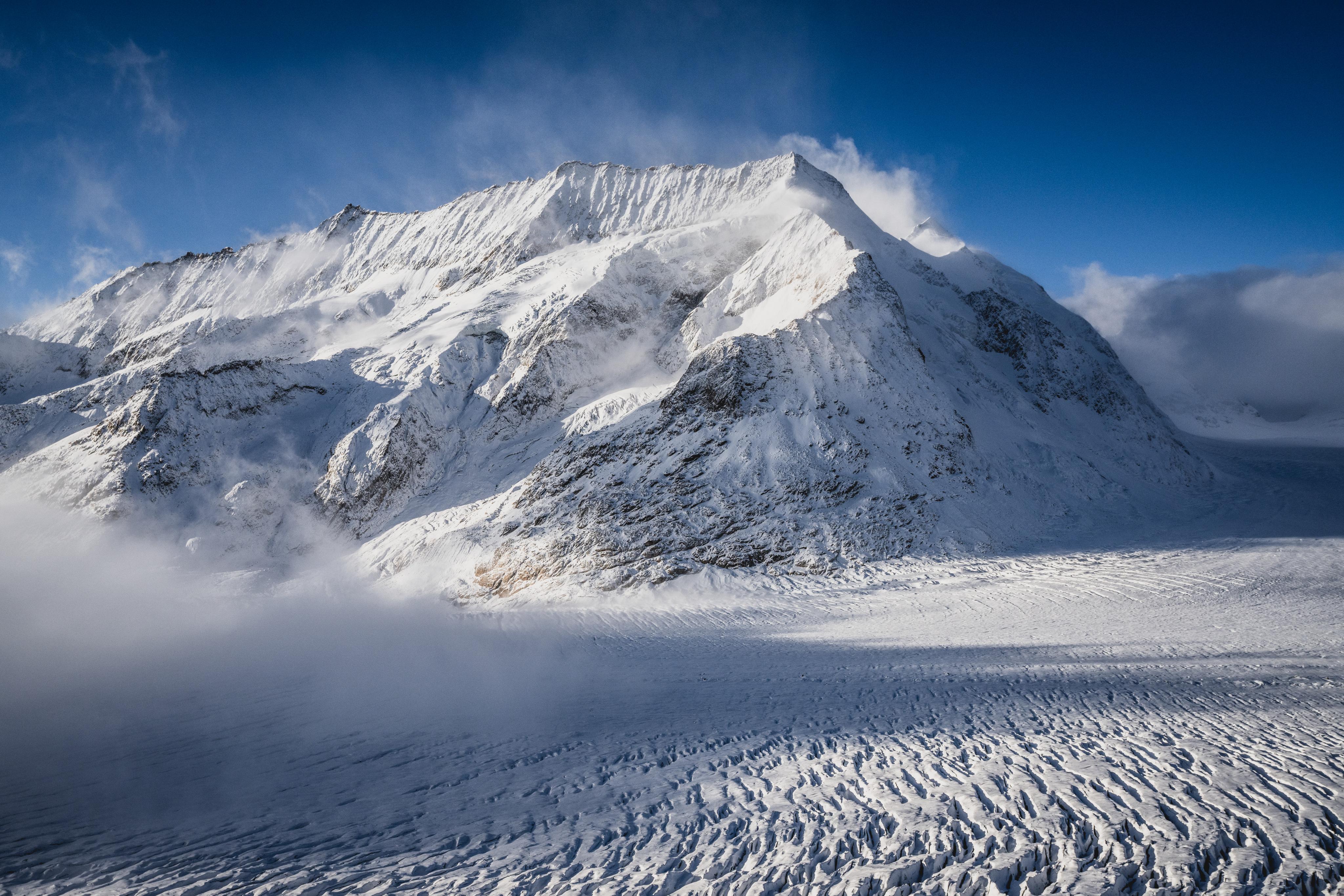 Snow-covered mountain peak with mist and clear blue sky in the background. The foreground showcases a textured glacier, perfect for Mammut mountaineering gear testing.