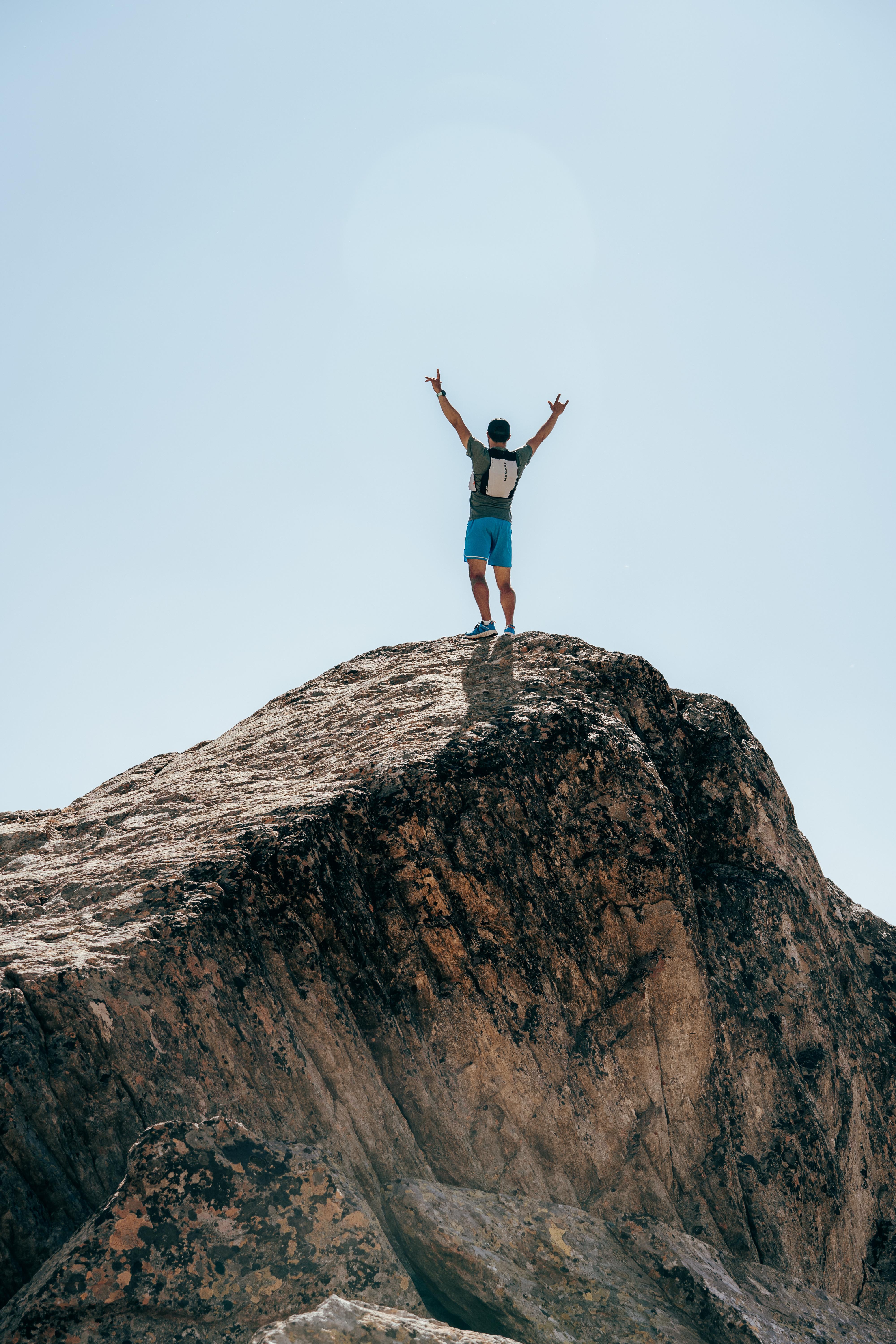 Person standing triumphantly on a rocky peak, wearing Mammut gear with arms raised under a clear blue sky.